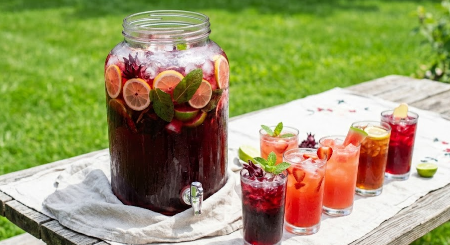 Juneteenth red drink recipes served outdoors — a large glass beverage dispenser filled with deep red hibiscus punch surrounded by five individual glasses of red drinks including strawberry lemonade, watermelon agua fresca, and sorrel punch on a wood picnic table with green grass in the background.